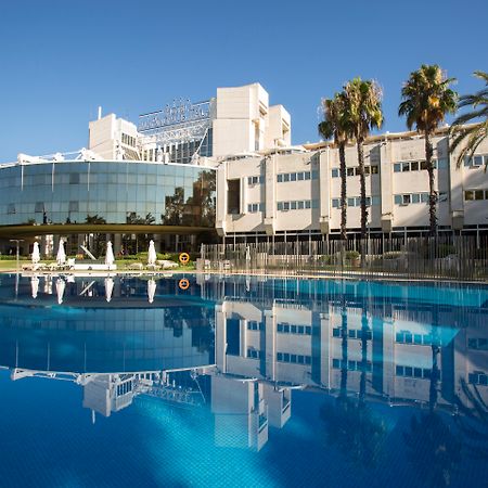 Silken Al-Andalus Palace Sevilla Exterior foto The photo features a modern hotel with a large glass facade reflecting in a calm blue swimming pool. The building is surrounded by palm trees, indicating a tropical or warm climate. The clear blue sky adds to the serene atmosphere, while lounge chair