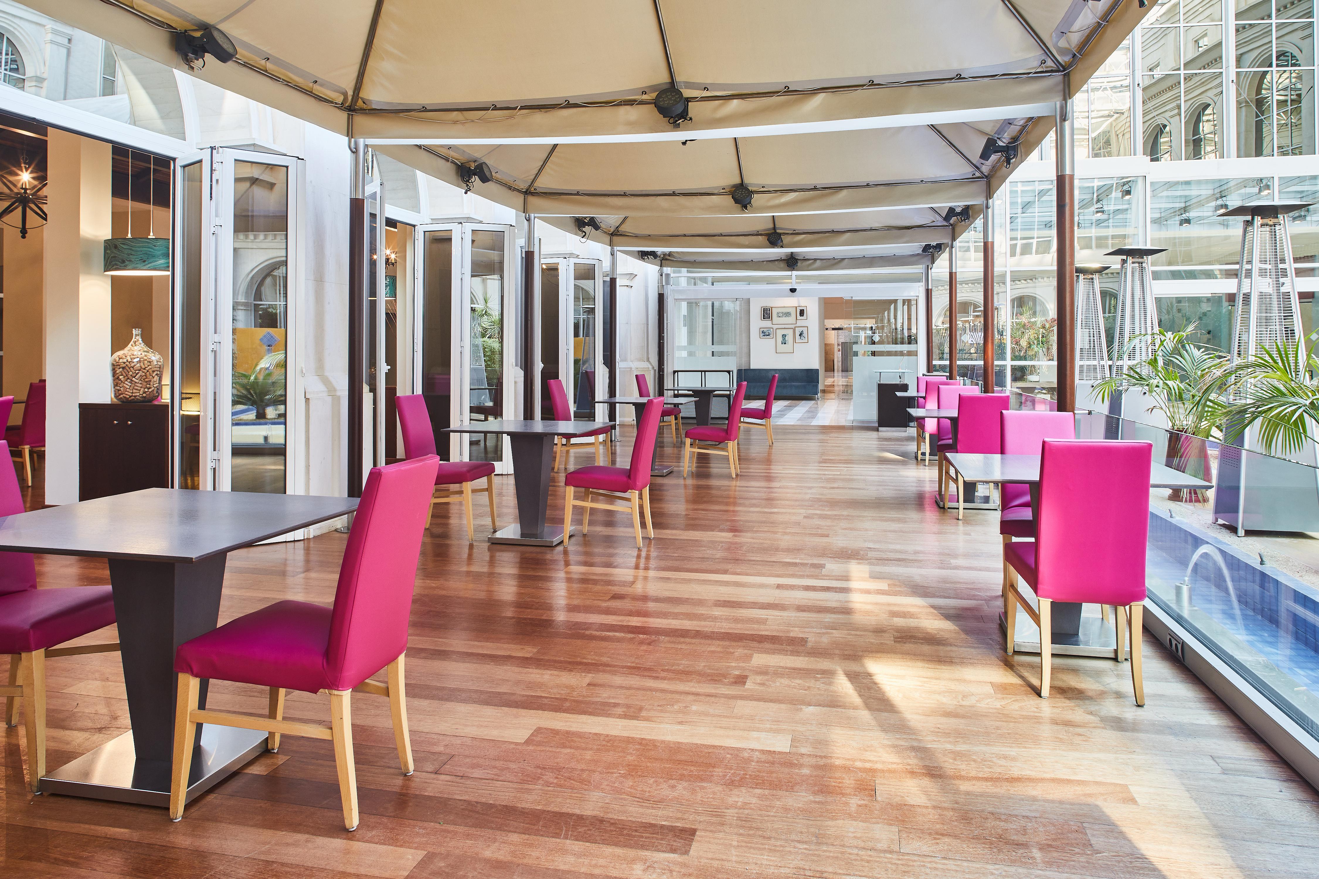 Silken Al-Andalus Palace Sevilla Exterior foto The photo shows a modern dining area with a sleek design. It features wooden flooring and tables set up in a row. Each table has a single pink chair positioned beside it. Large glass windows or doors line one side, creating an open and airy atmospher