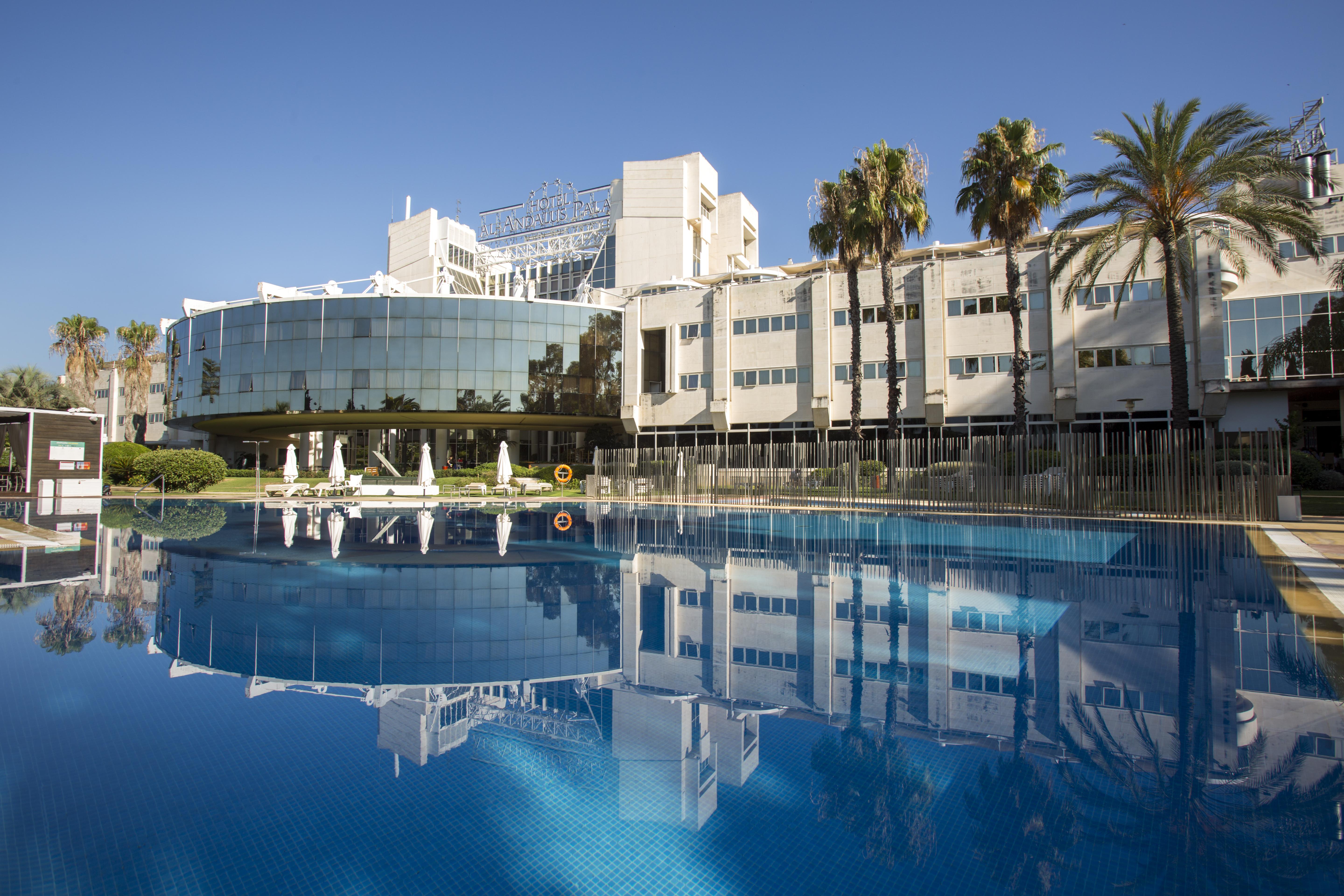 Silken Al-Andalus Palace Sevilla Exterior foto The photo features a modern hotel with a large glass facade reflecting in a calm blue swimming pool. The building is surrounded by palm trees, indicating a tropical or warm climate. The clear blue sky adds to the serene atmosphere, while lounge chair
